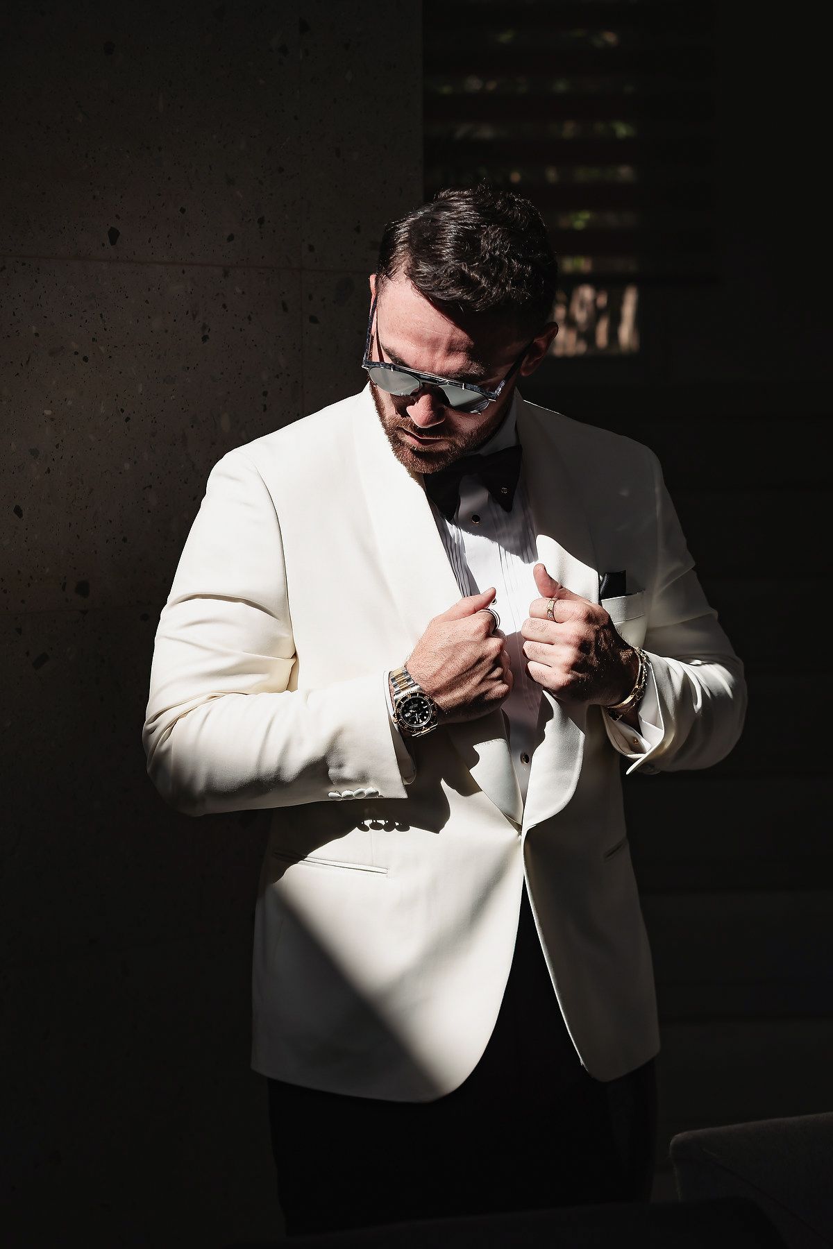 Stylish man in white tuxedo jacket and bow tie adjusting his suit, wearing sunglasses and a watch.