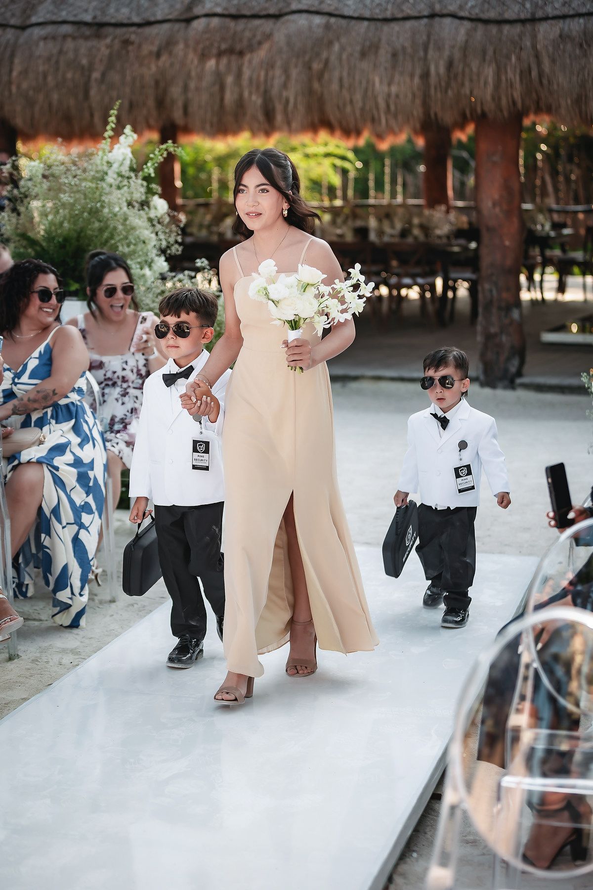 Bridesmaid in champagne gown walks down aisle with two young ring bearers in tuxedos at outdoor wedding.