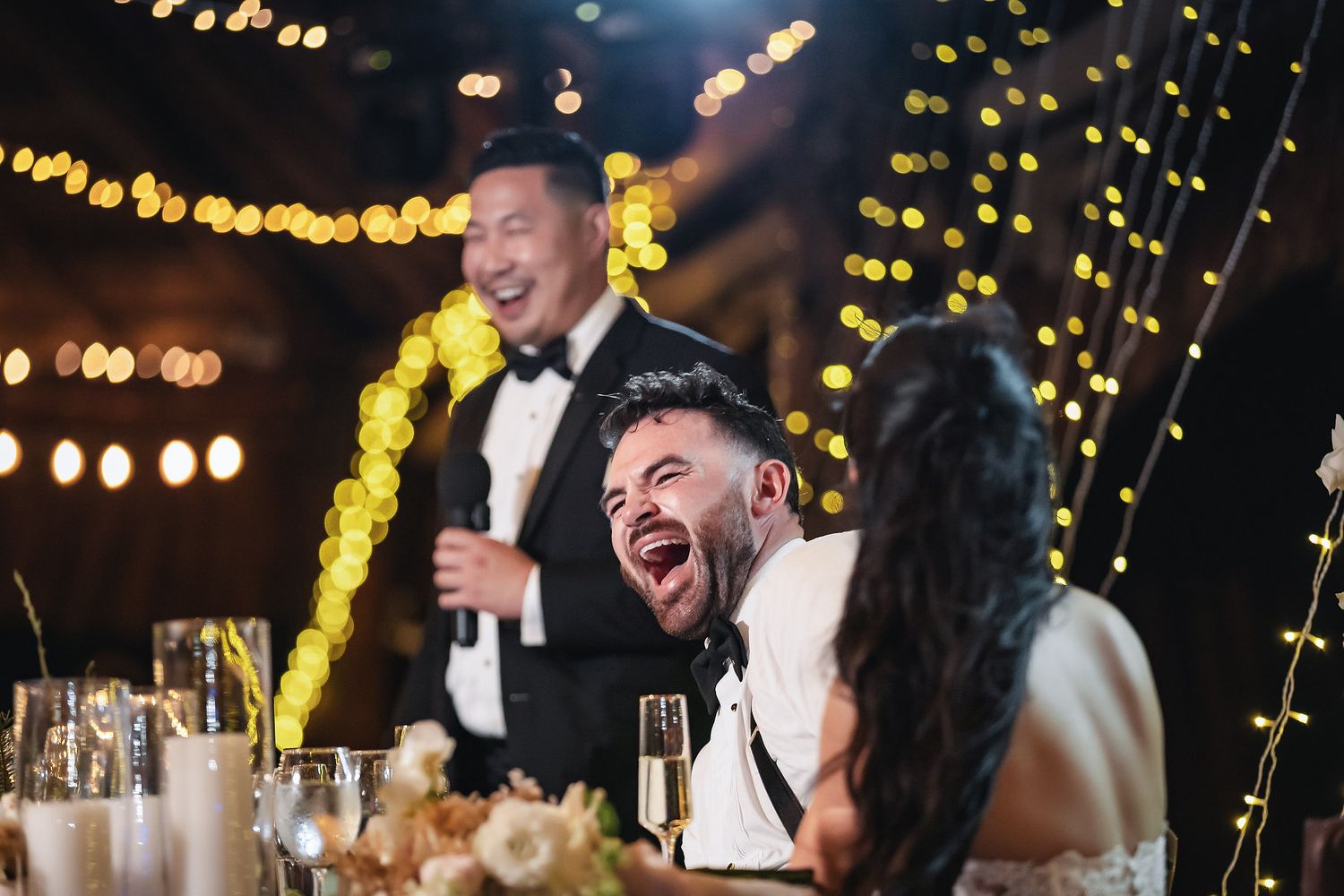 Groom laughs joyfully during wedding reception speech, surrounded by guests and warm string lights.
