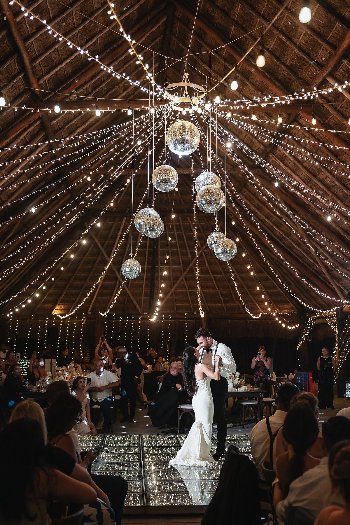 Bride and groom share first dance under disco balls and fairy lights in a romantic barn wedding venue.