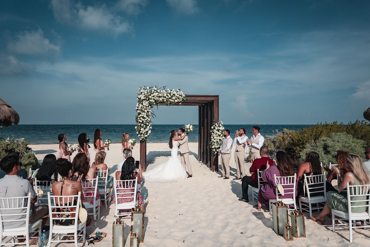 Romantic beach wedding ceremony with couple kissing under floral arch, guests seated on white chairs, ocean backdrop.