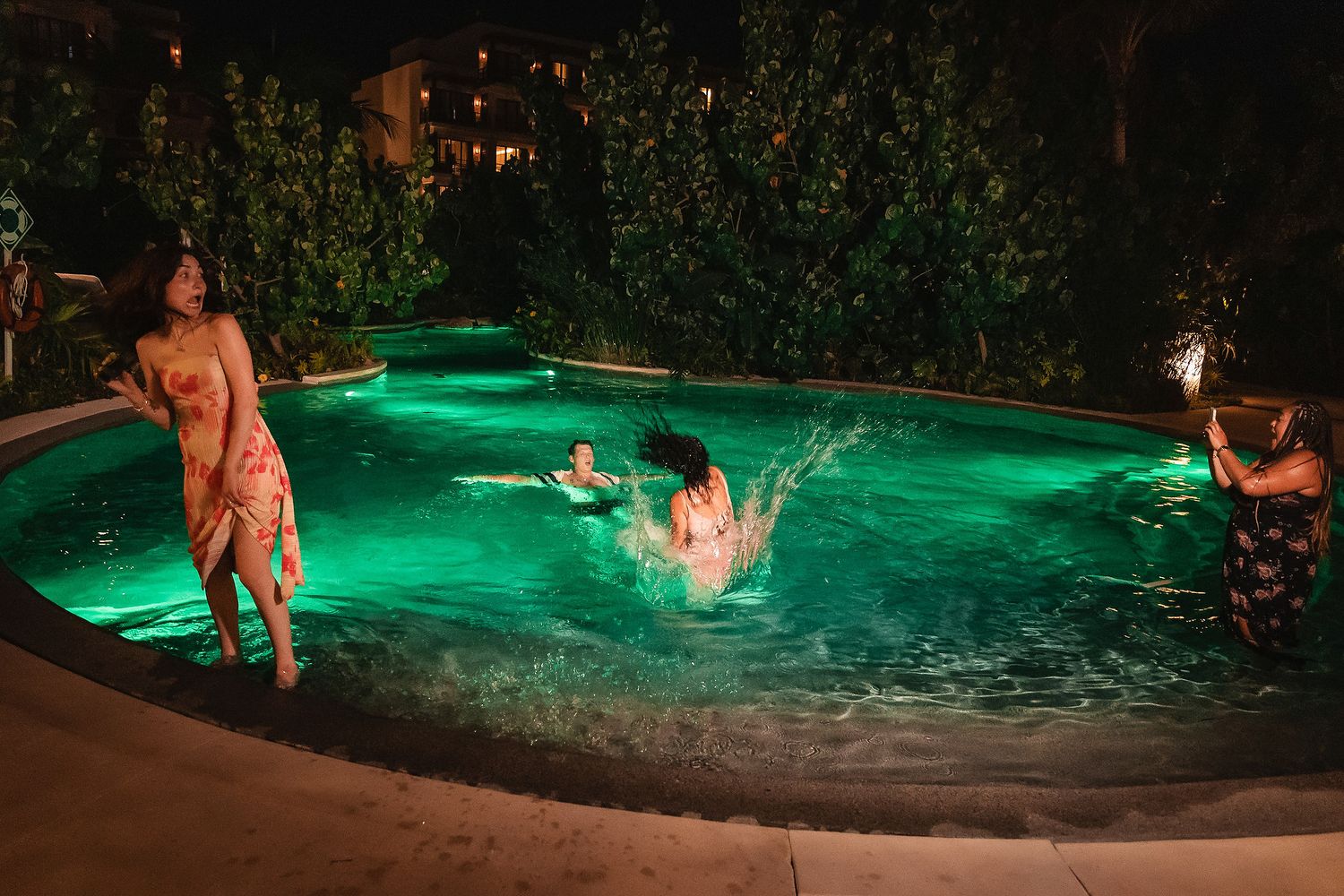Friends enjoying a nighttime pool party with green underwater lights and tropical surroundings.