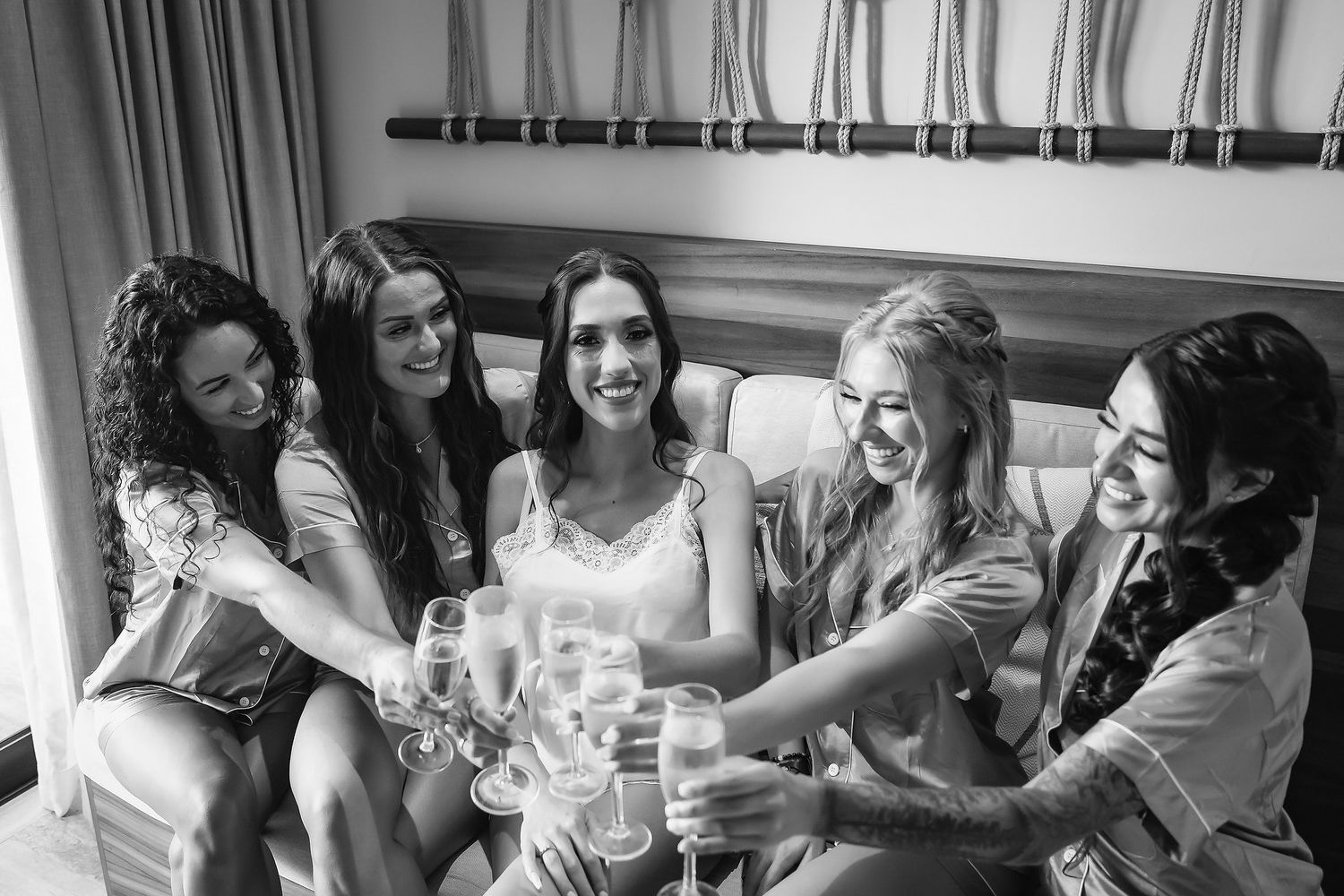 Black and white photo of a bride and bridesmaids in robes toasting with champagne glasses while seated together.