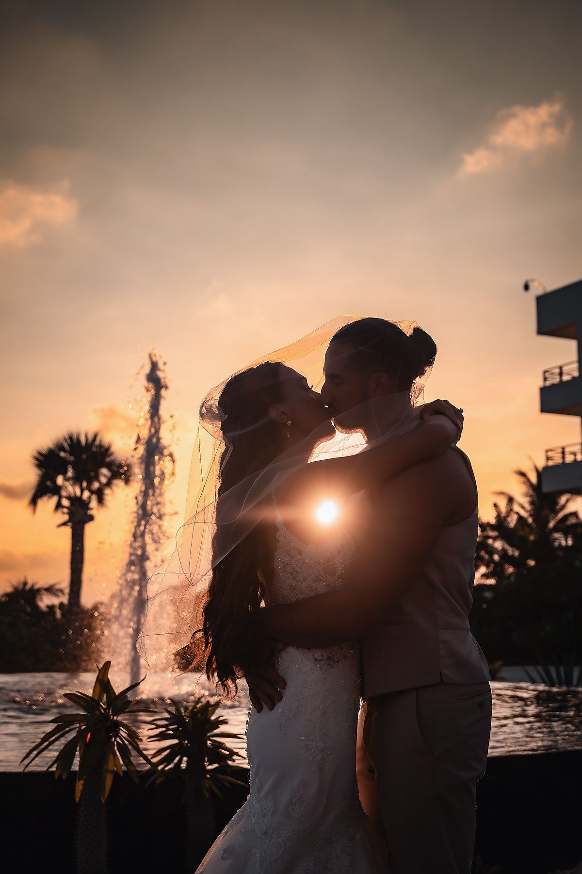 Silhouette of bride and groom kissing at sunset near a fountain, with palm trees and golden sky backdrop.