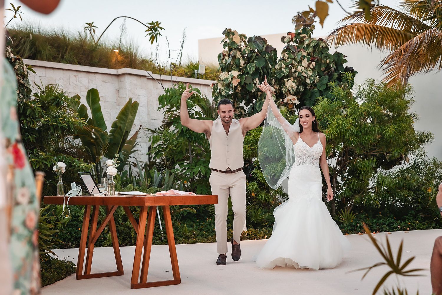 Joyful newlyweds celebrate after wedding ceremony outdoors, groom raising arms in excitement beside bride in white gown.