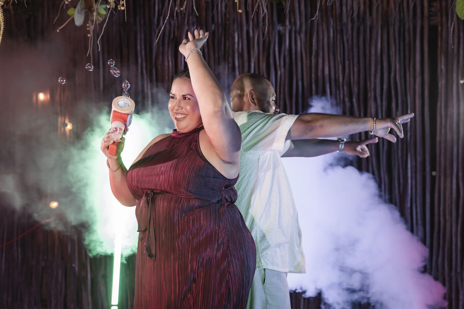Two plus-size women dancing at a party with colorful smoke, one holding a drink under festive lights.