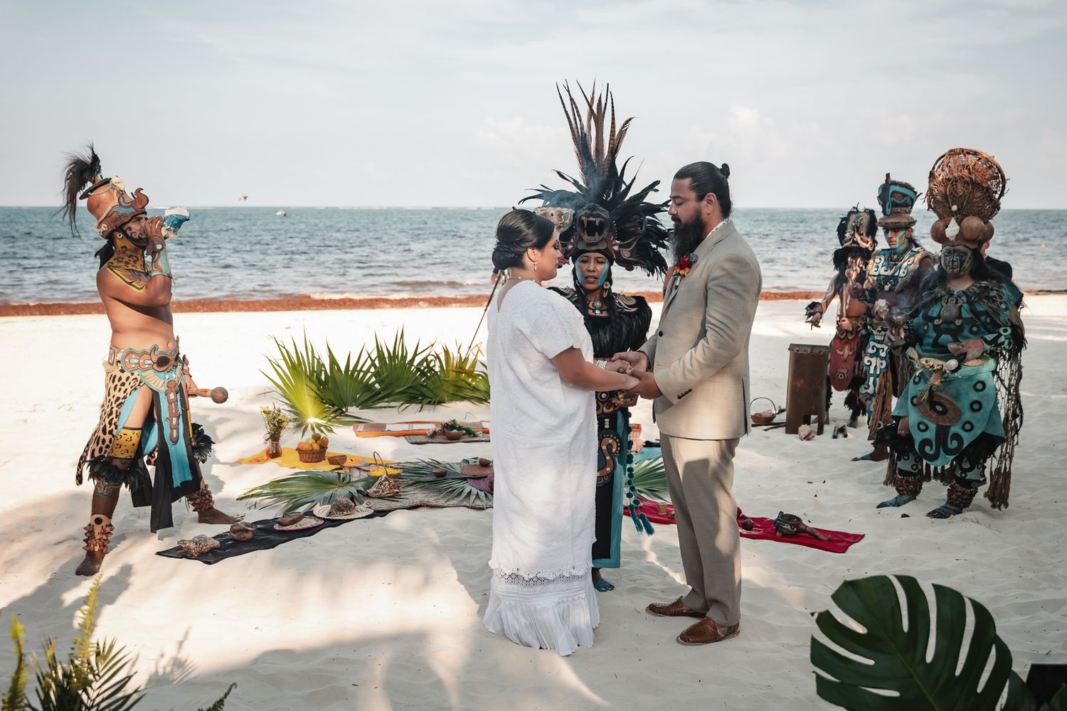 Couple exchanges vows on beach surrounded by Mayan performers in traditional ceremonial attire.