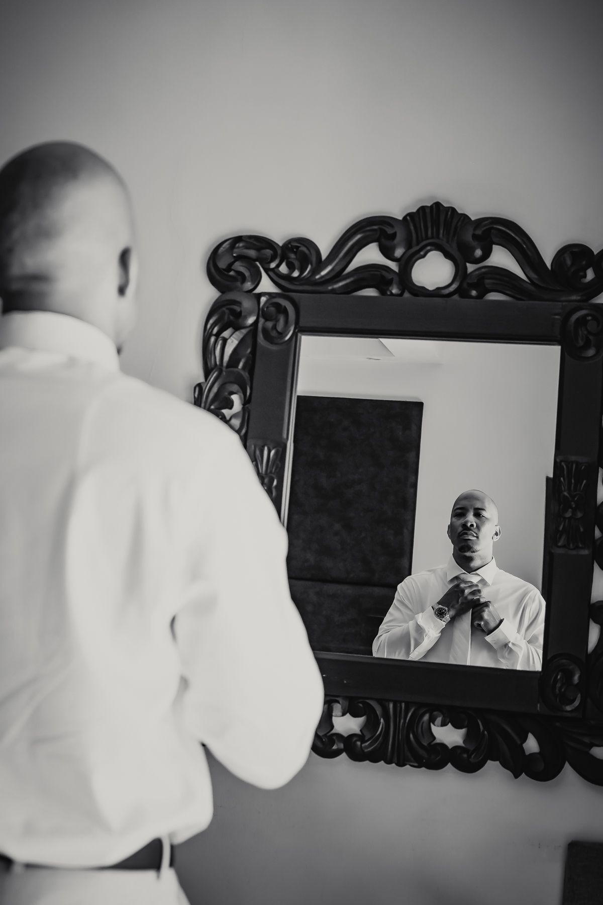 Groom adjusting his tie while looking into an ornate black mirror in black and white photo.