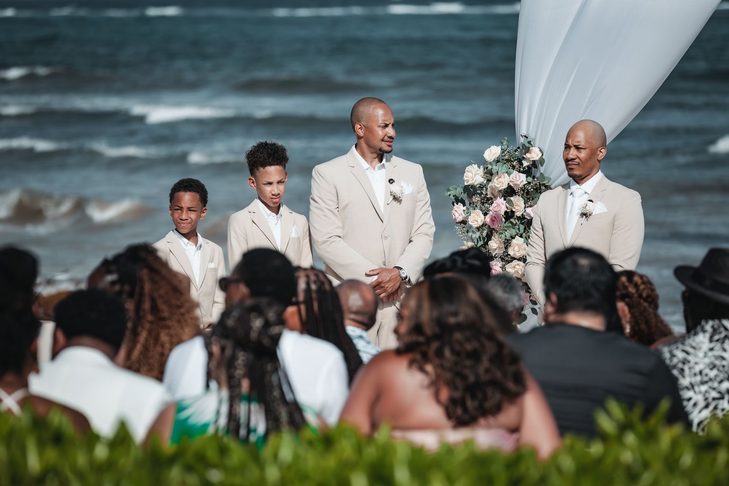 Groom and groomsmen in cream suits stand at beach wedding altar with floral arrangement and ocean backdrop.