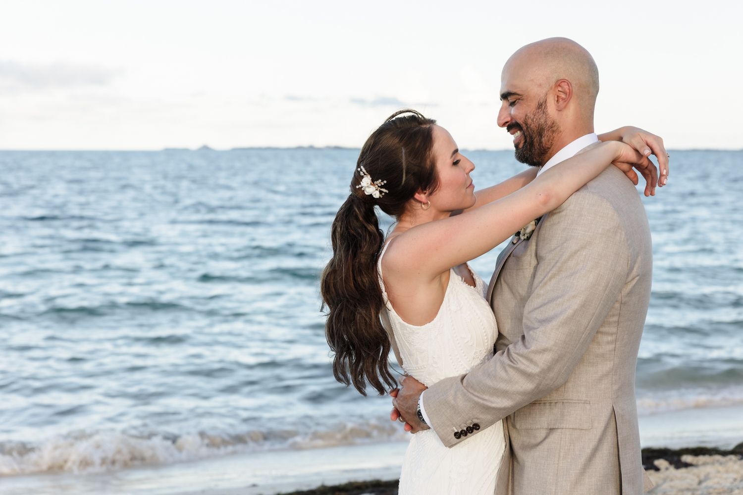 Newlywed couple embracing on a beach at sunset, bride in white dress with floral hair accessory, groom in tan suit.
