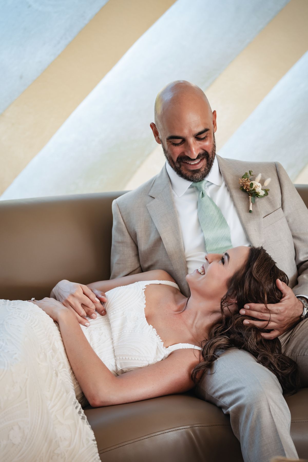 Bride and groom share a tender moment on a leather sofa, with the groom cradling her head and smiling warmly.
