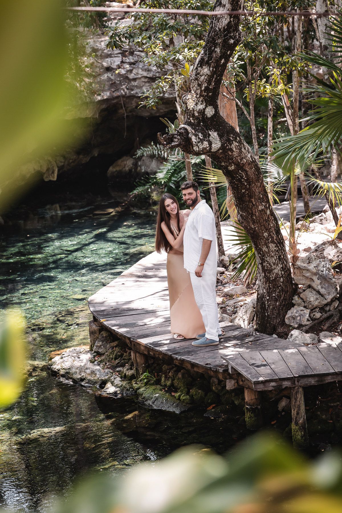 Couple embracing on wooden dock beside turquoise cenote waters surrounded by lush tropical jungle greenery.