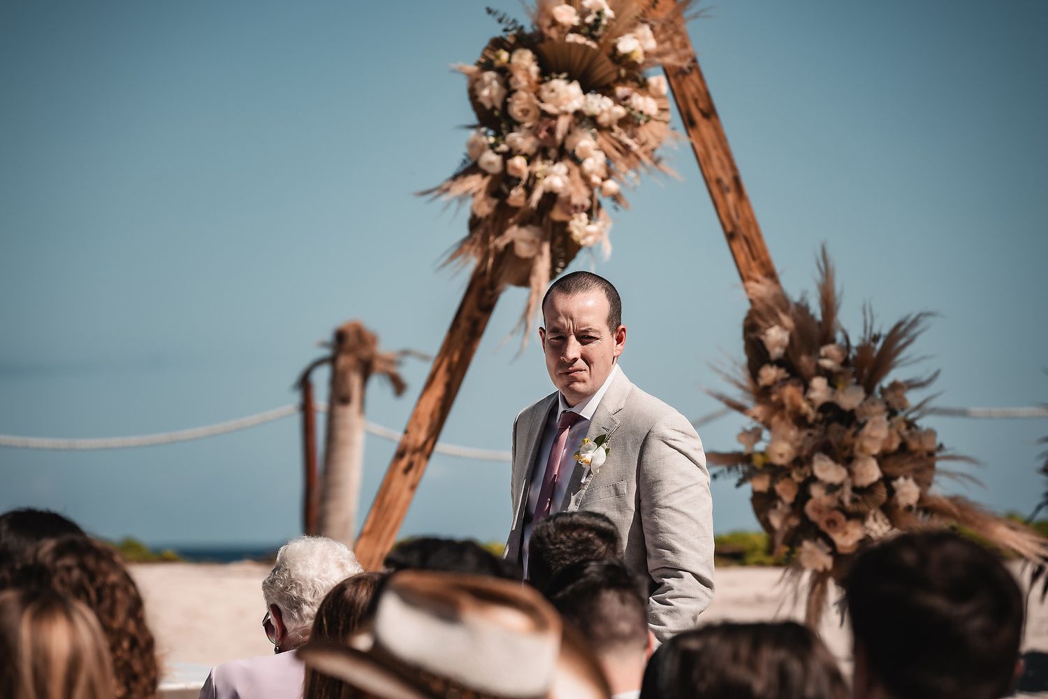 Outdoor beach wedding ceremony setup with palm trees and wooden arch against blue sky.