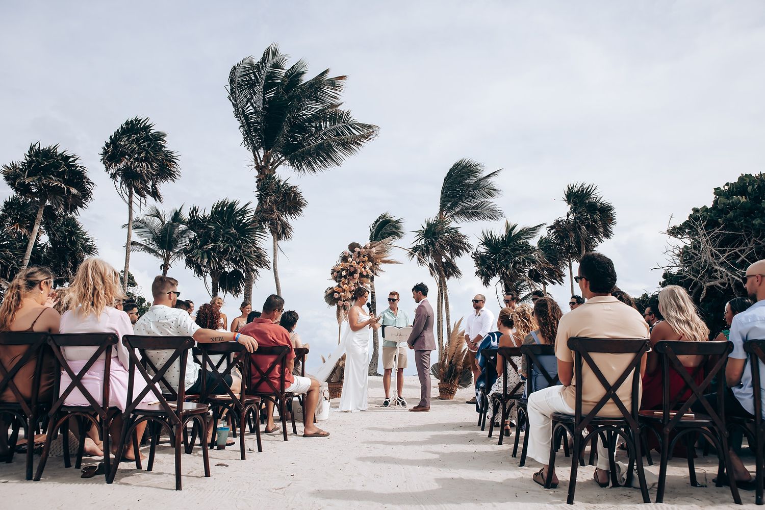 Panoramic view of palm trees swaying in wind along a beachfront wedding ceremony setup with wooden chairs.