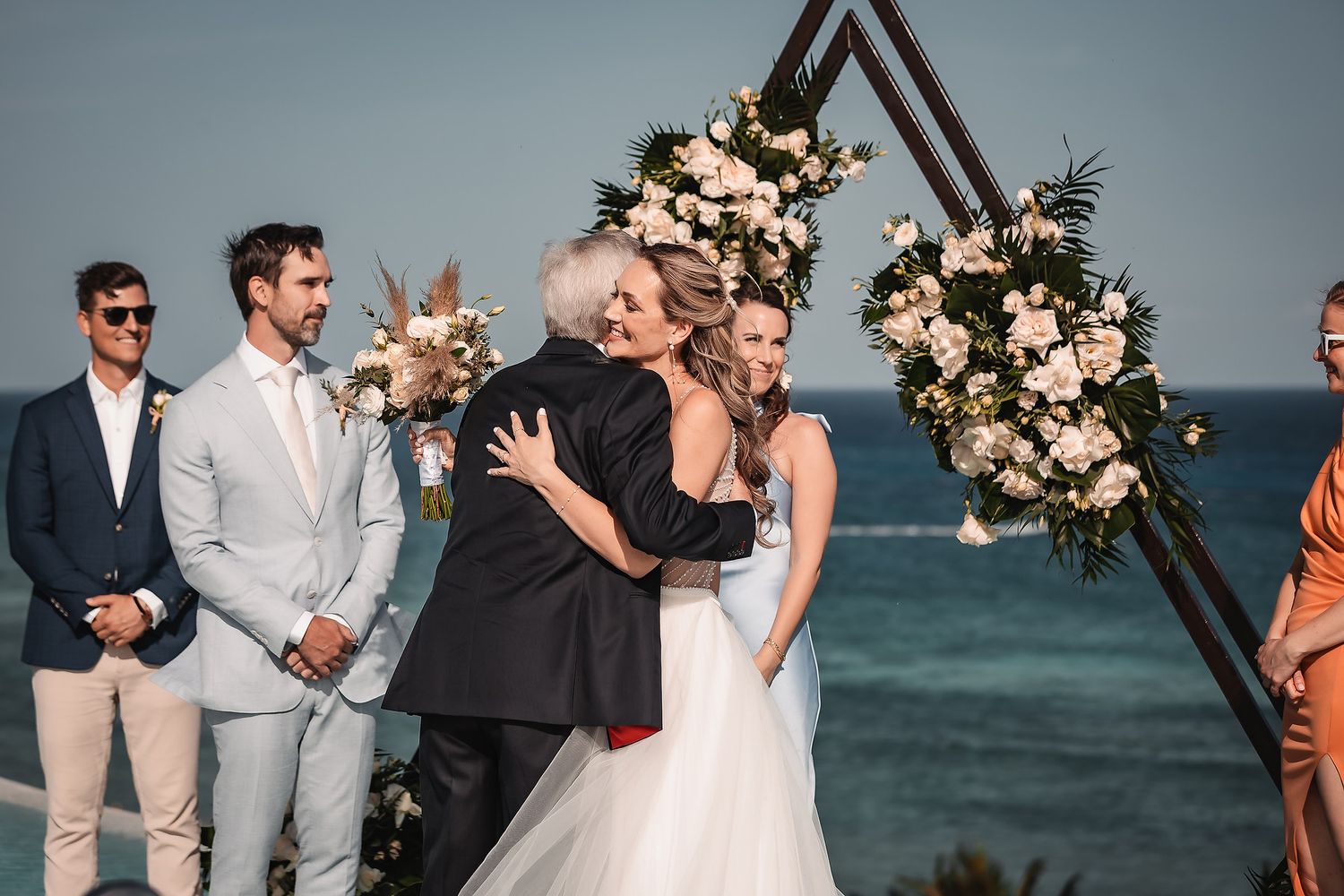 Beachfront wedding ceremony under a triangular arch decorated with blush and cream flowers.
