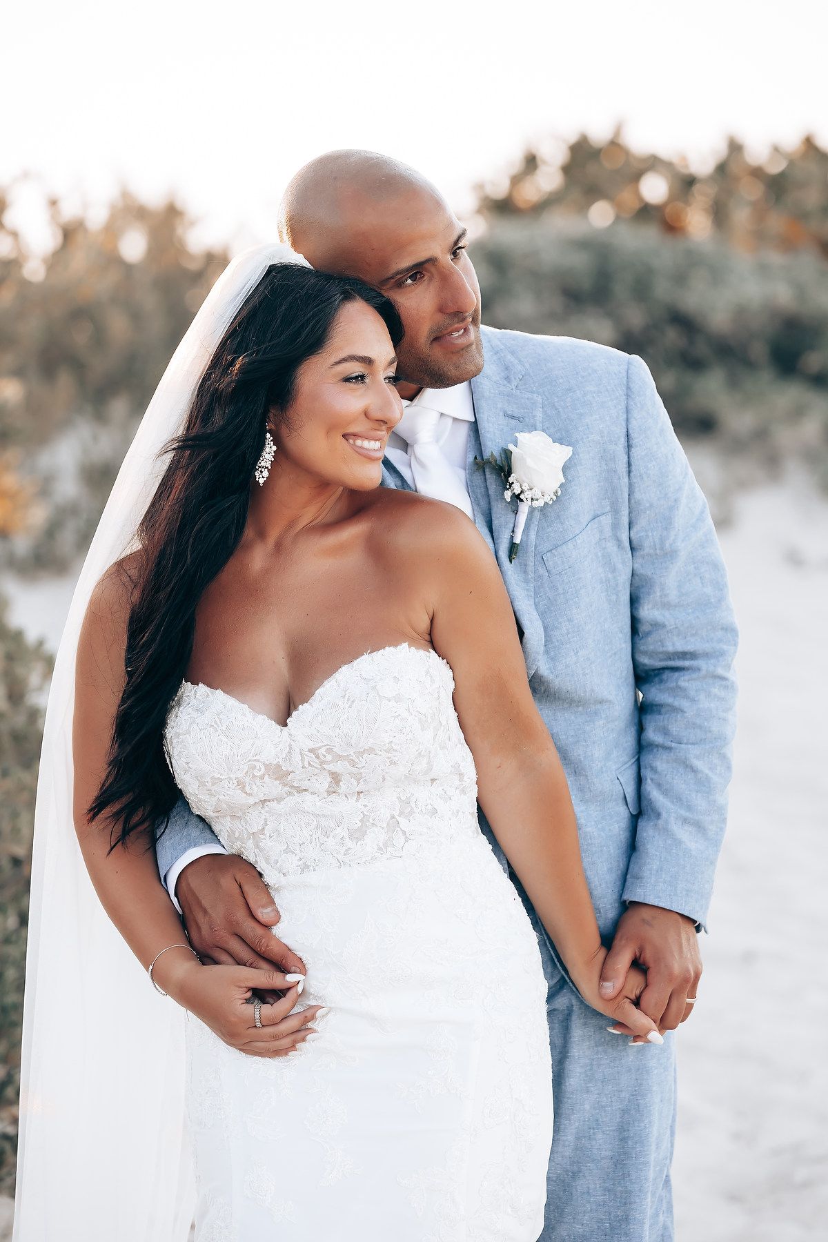 A romantic beach wedding photoshoot featuring a couple in a light blue suit and white lace dress at sunset.