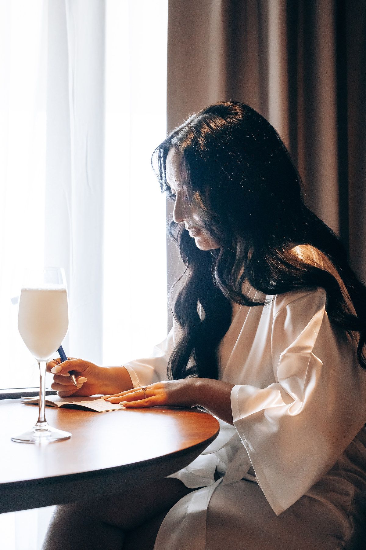 Woman in white sweater sits at table writing in notebook while enjoying a glass of wine near window.