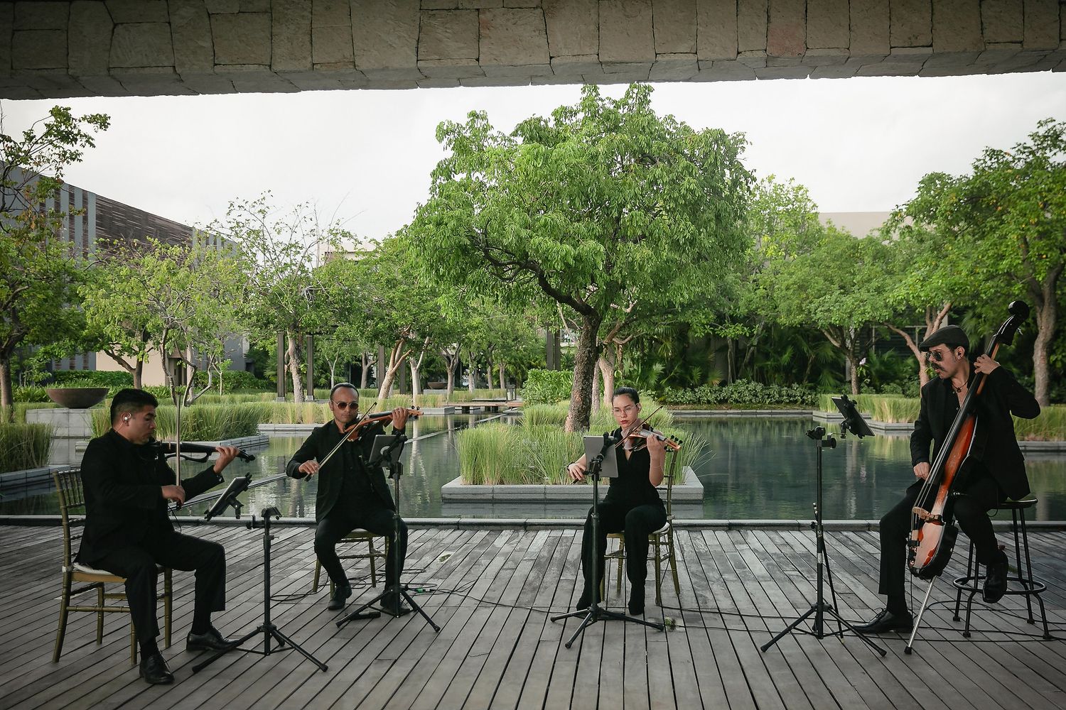 A string quartet performs outdoors on a covered deck overlooking a tranquil pond surrounded by lush greenery.