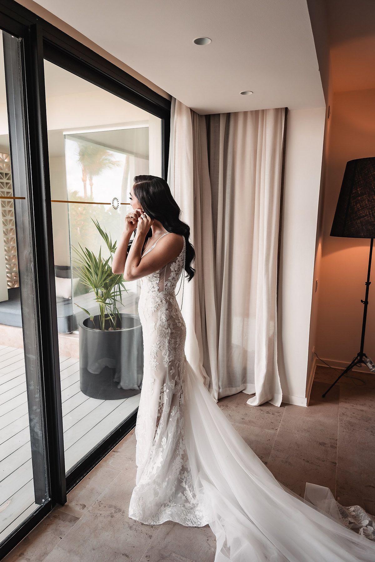 A bride in a flowing white lace gown stands in an elegant hallway with floor-to-ceiling windows and white curtains.