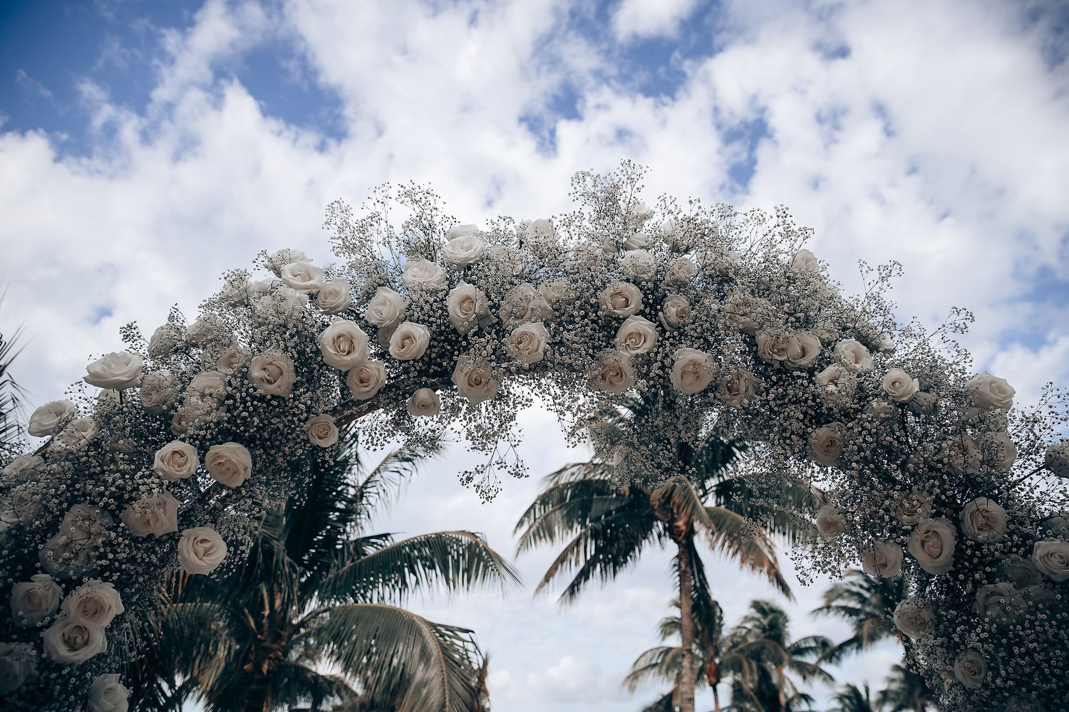 Palm trees and white floral arrangements create a tropical wedding ceremony backdrop under blue skies.