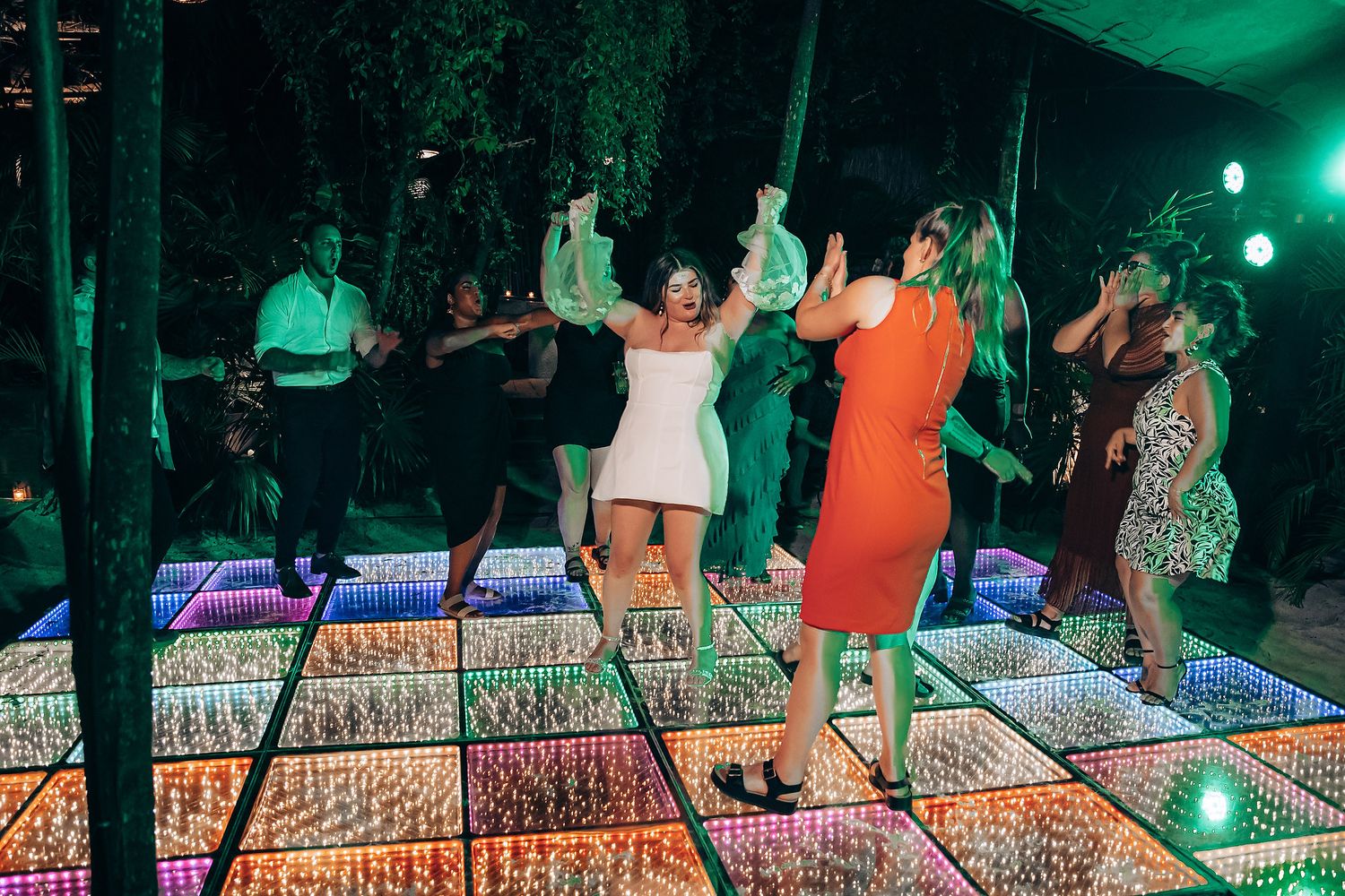 People dance on an illuminated LED dance floor with colorful green lighting at a party event.