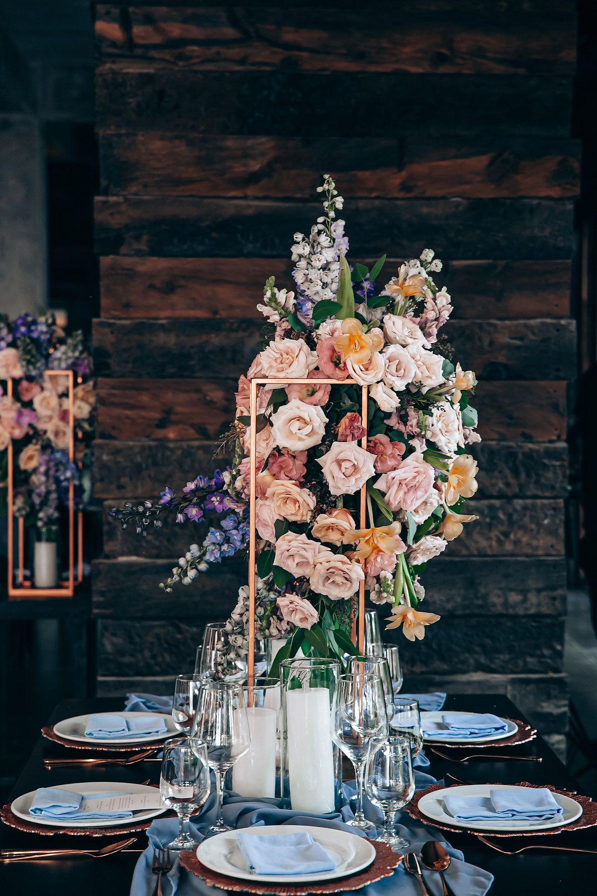 An elegant table setting with a tall floral centerpiece featuring pink roses and delicate blooms against dark wood walls.