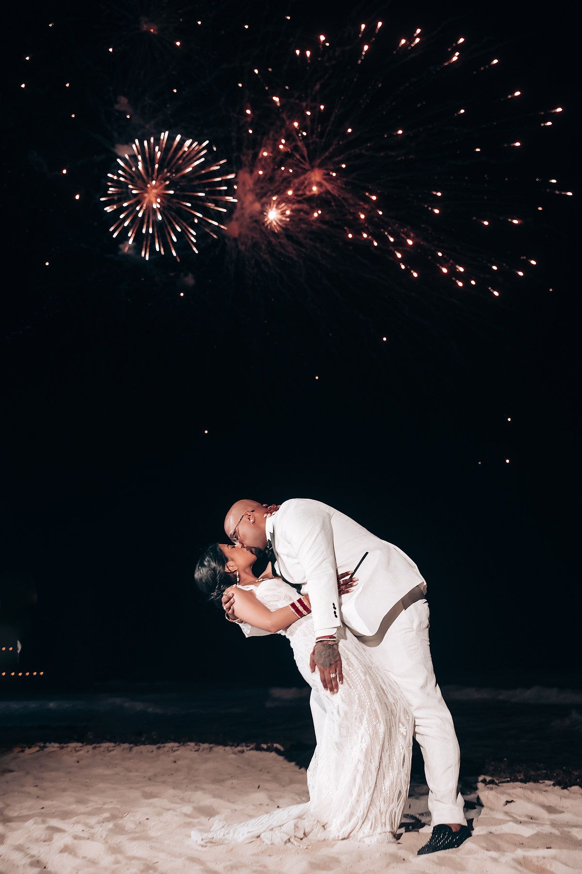 A romantic dip kiss under bursting red fireworks on a moonlit beach.