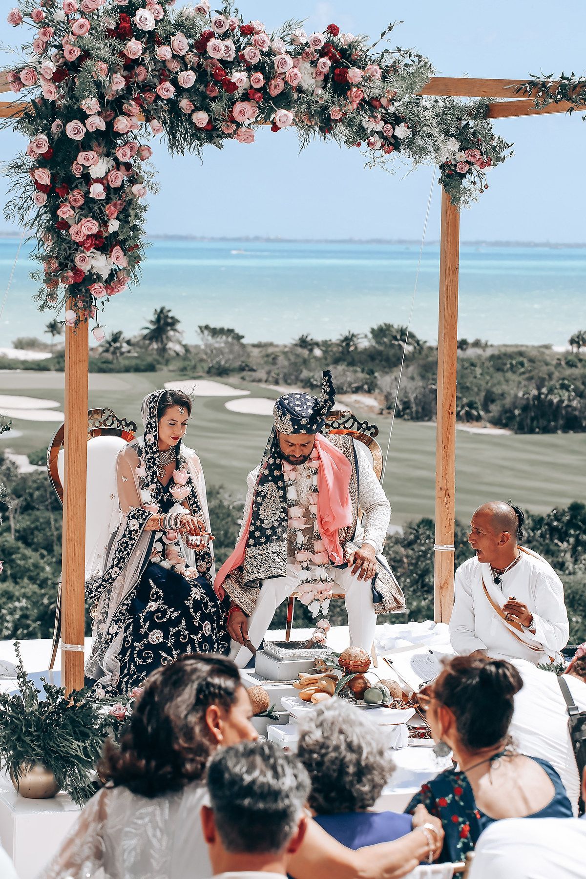 Wedding guests dine at decorated tables under a floral arch with an ocean view backdrop.