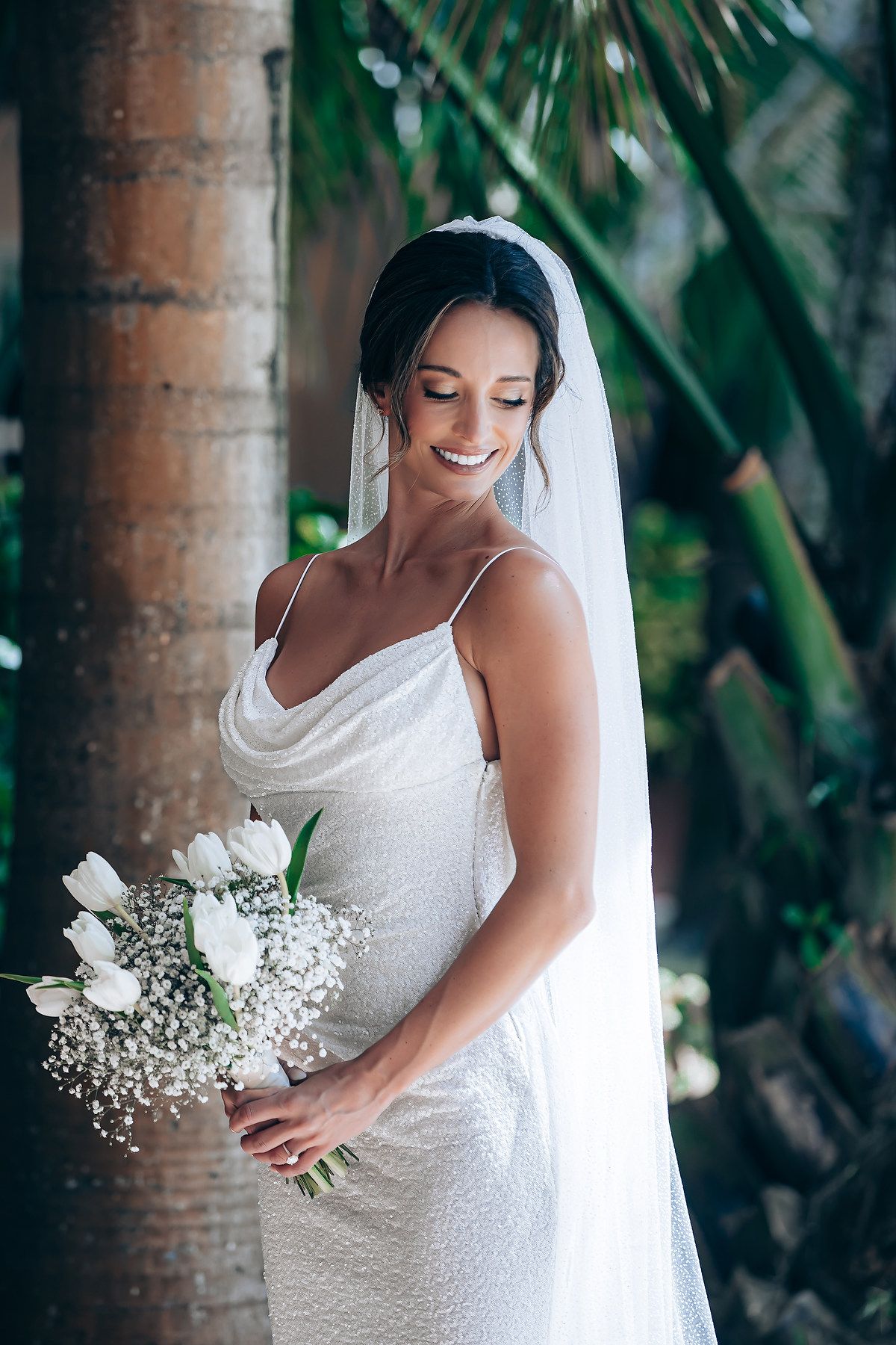 A bride in a white wedding dress holds a delicate bouquet of white flowers and baby's breath against a tropical backdrop.