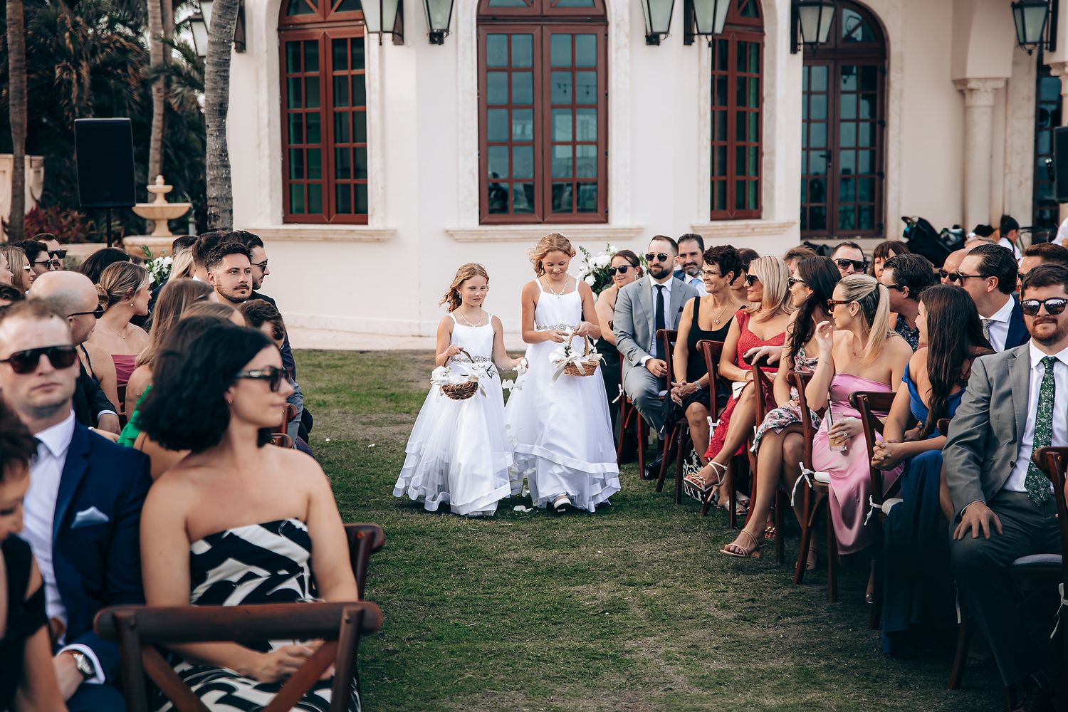 Wedding guests sit on white chairs on a lawn watching a ceremony in front of a Mediterranean-style building.