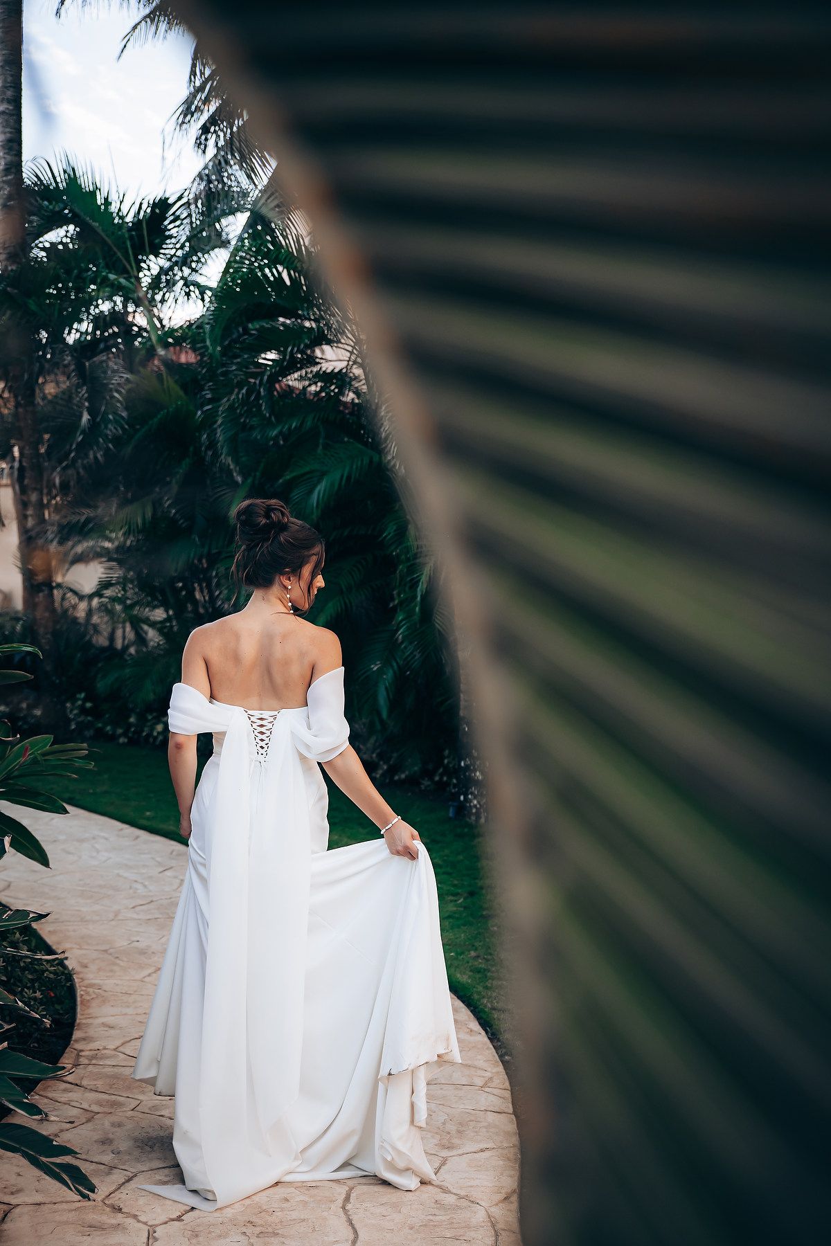 Bride in flowing white wedding dress sits on stone steps with palm trees in background at sunset.