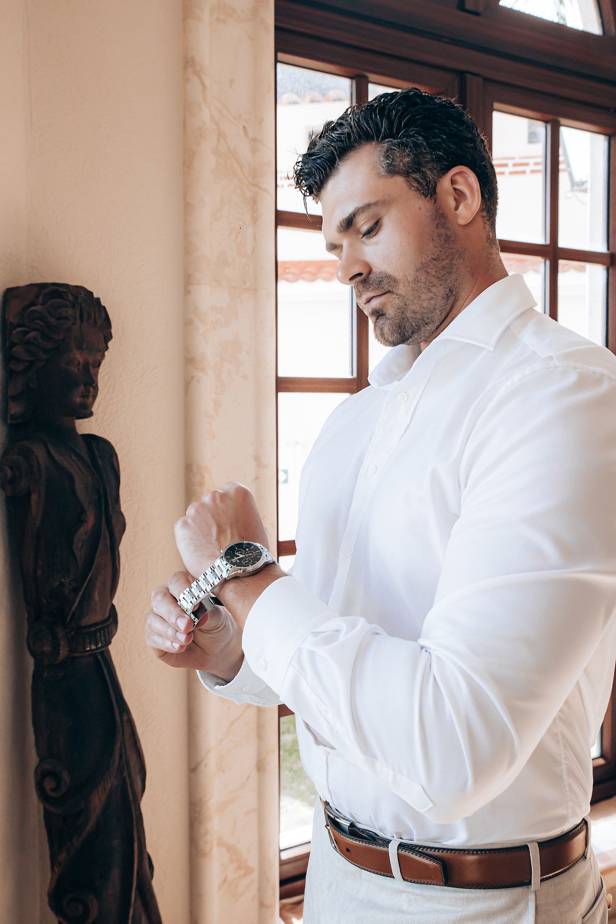 Man in white dress shirt adjusts his cufflinks while getting dressed in formal attire near a window.