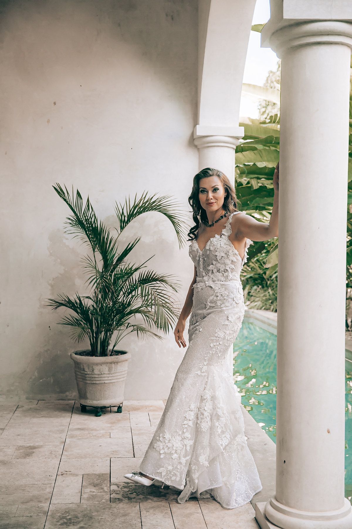 A bride in a white lace wedding dress poses elegantly between classical white columns with a potted palm plant nearby.
