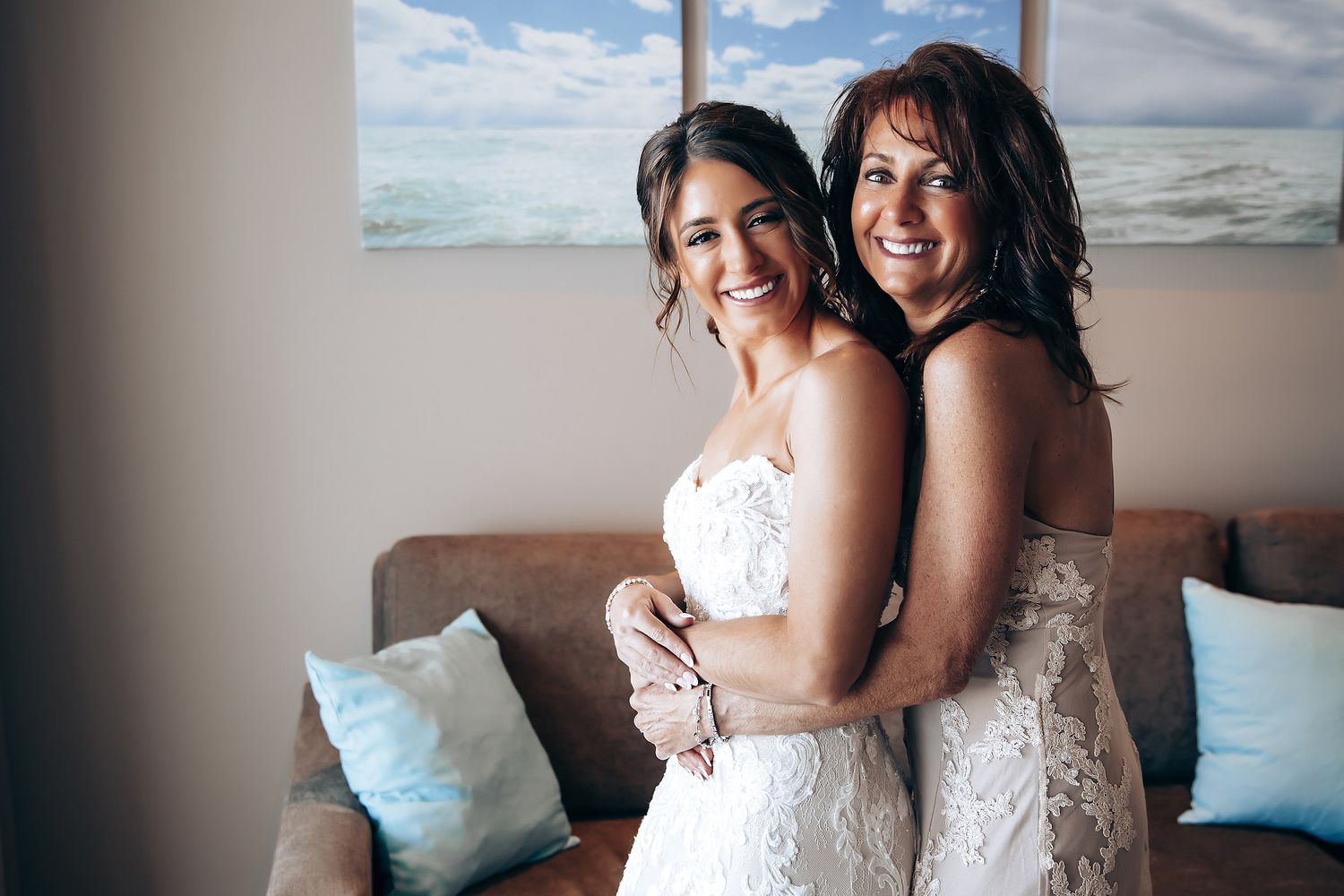 Two people in white dresses embrace and smile while sitting on a couch with blue pillows.