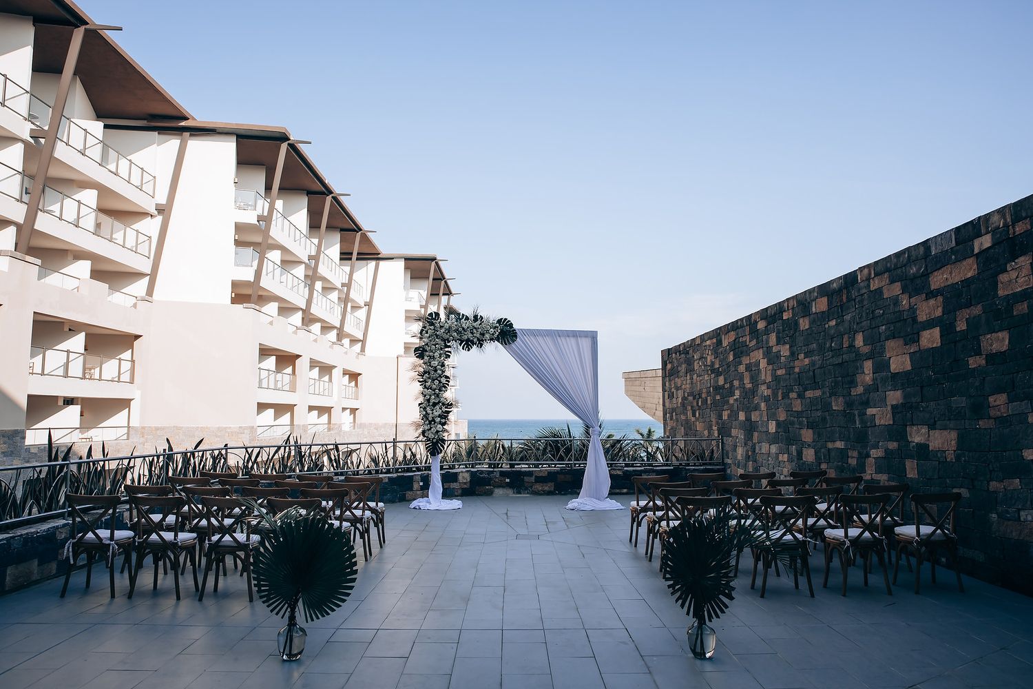 Wedding ceremony setup with white chairs and floral arrangements against a modern building backdrop.