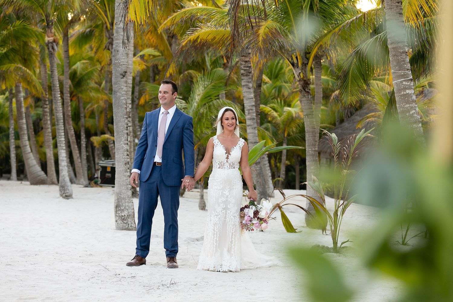 Newlyweds walk hand in hand along a sandy beach path lined with lush palm trees in a tropical setting.