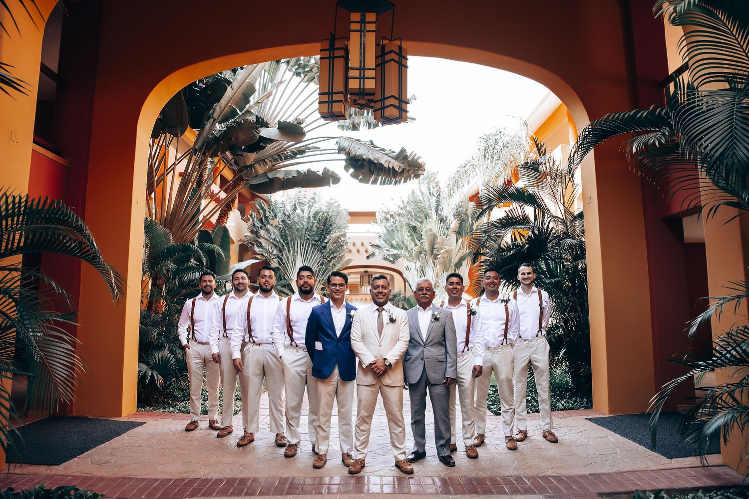 Groomsmen in white and navy attire pose under an elegant arched walkway with tropical landscaping.
