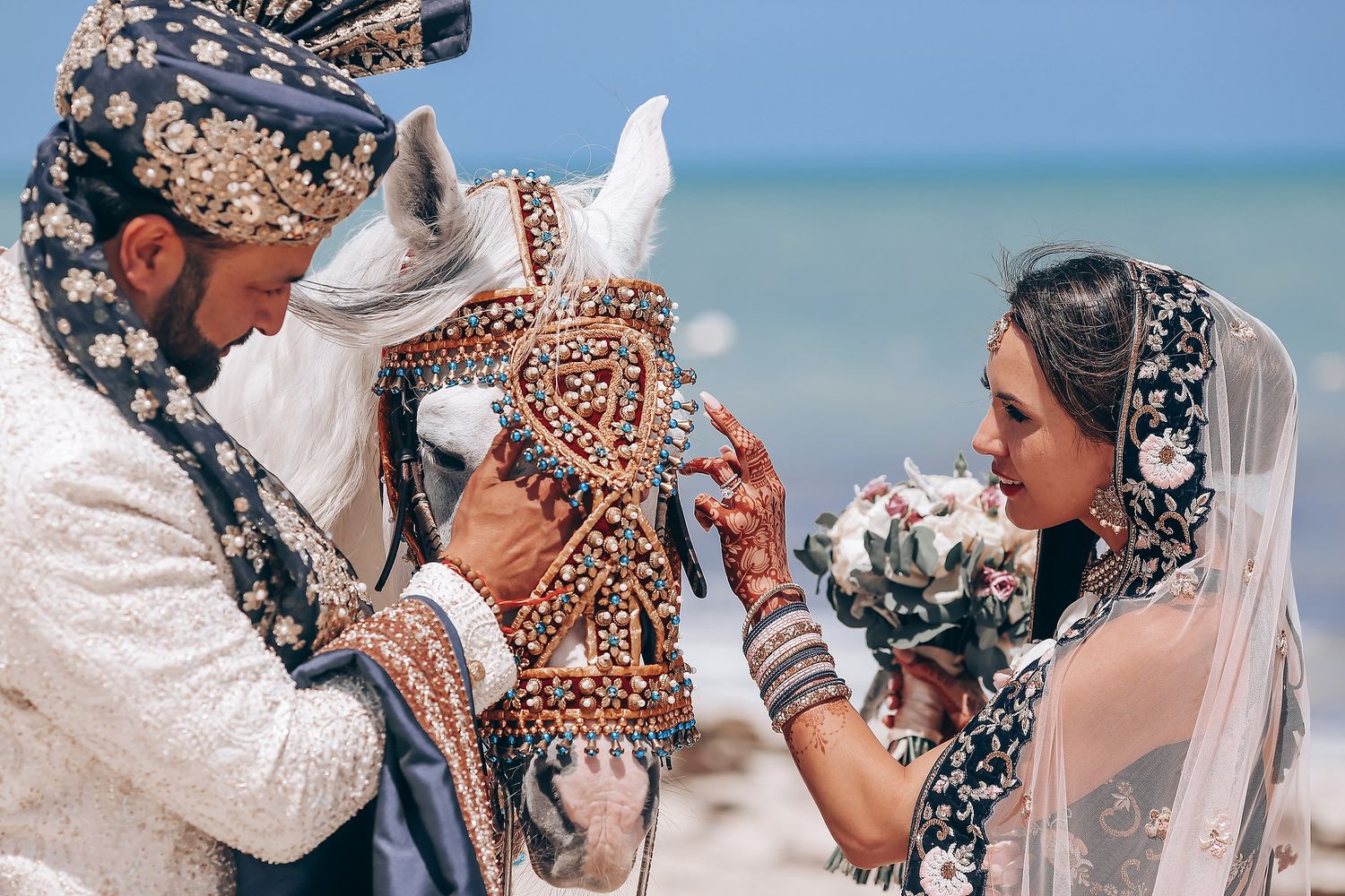 A group of people in traditional wedding attire embrace during a ceremony with confetti falling on a beach.