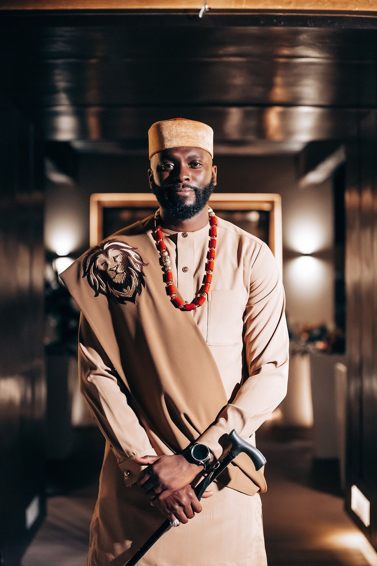 A person in a beige suit and red beaded necklace poses dramatically in a dimly lit hallway setting.