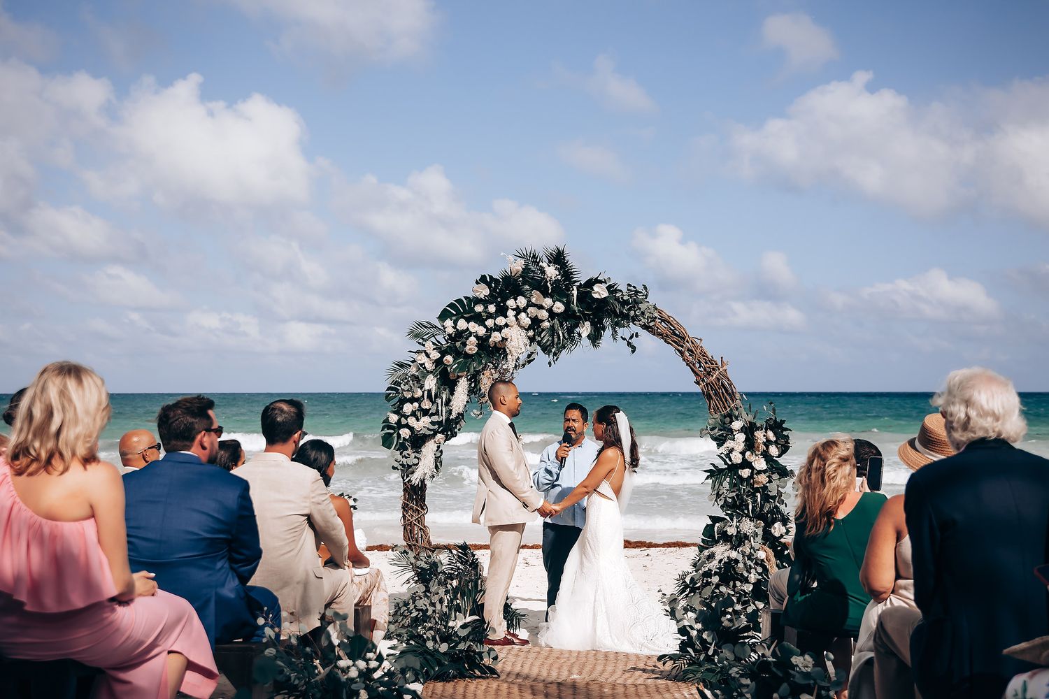 Panoramic view of a beachside wedding with guests gathered around a decorated arch.