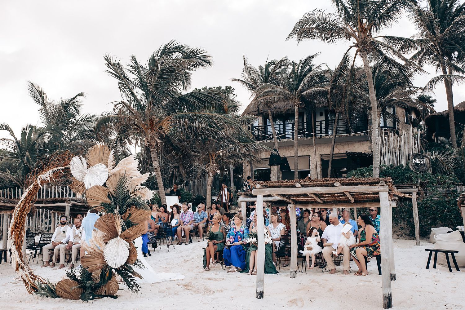 Wide angle shot of tropical beach wedding ceremony with palm trees and seated guests.