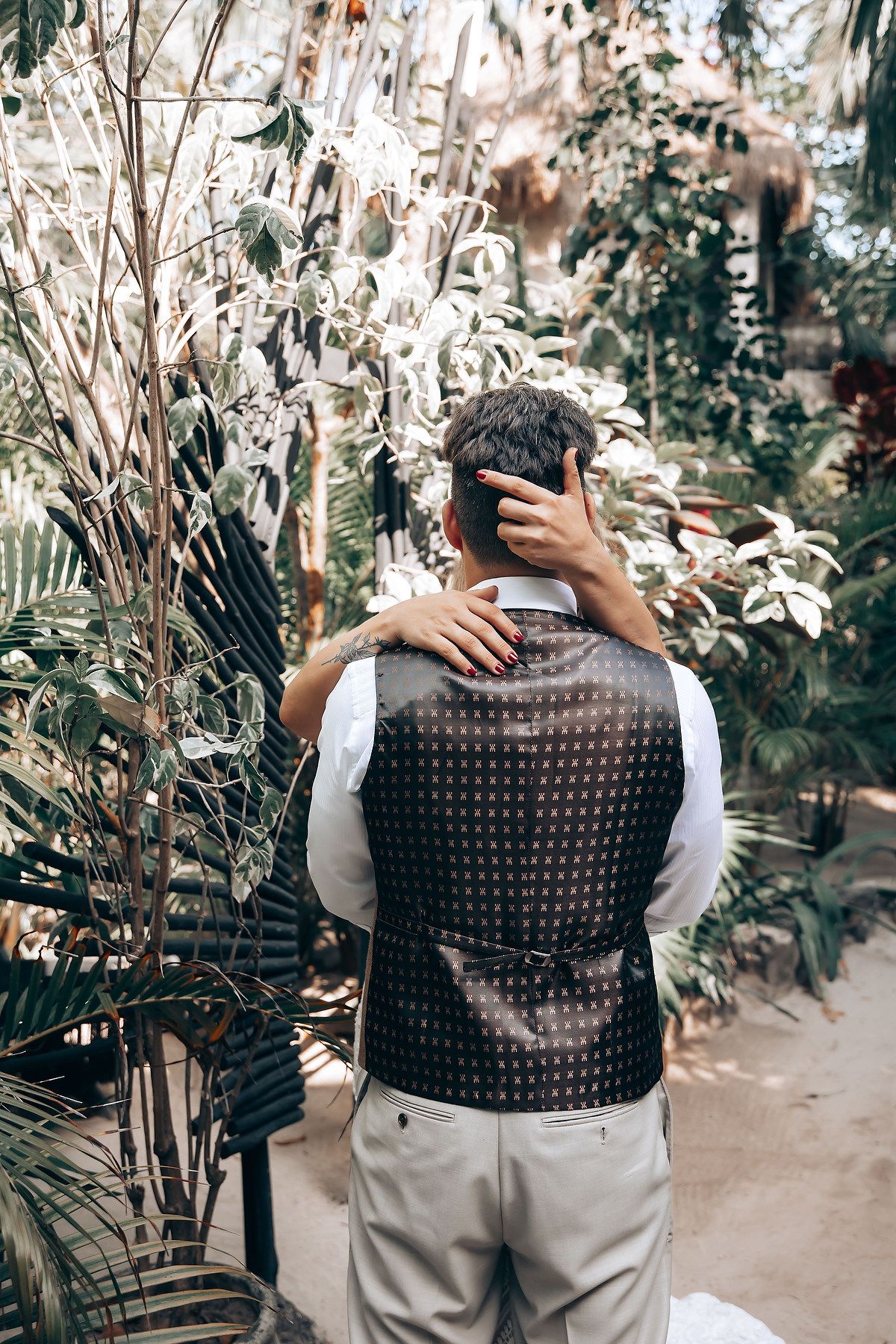 Back view of groom wearing patterned vest against tropical garden background.