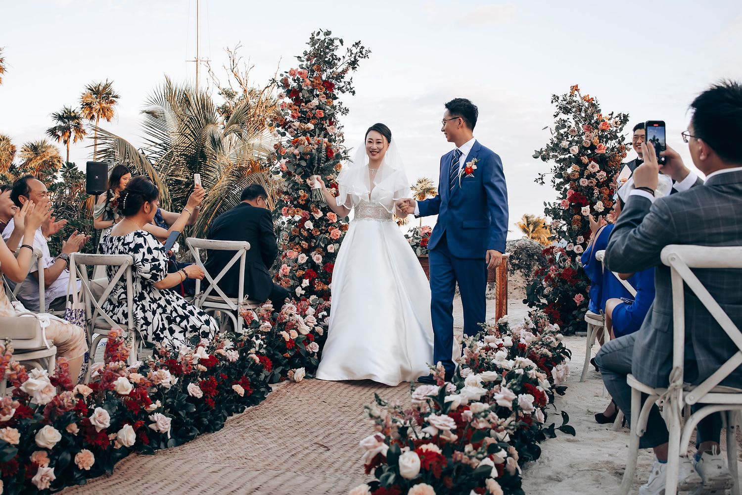 Wedding ceremony on a beachfront deck with pink and red floral arrangements.