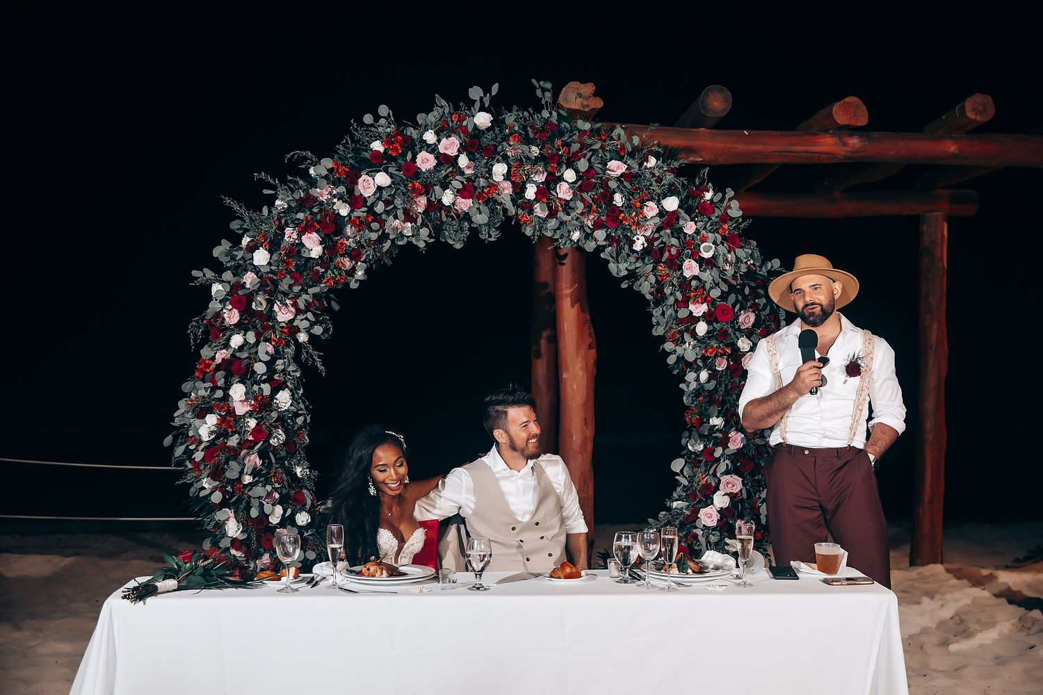 Wedding reception setup with floral arch and white tablecloths at night.