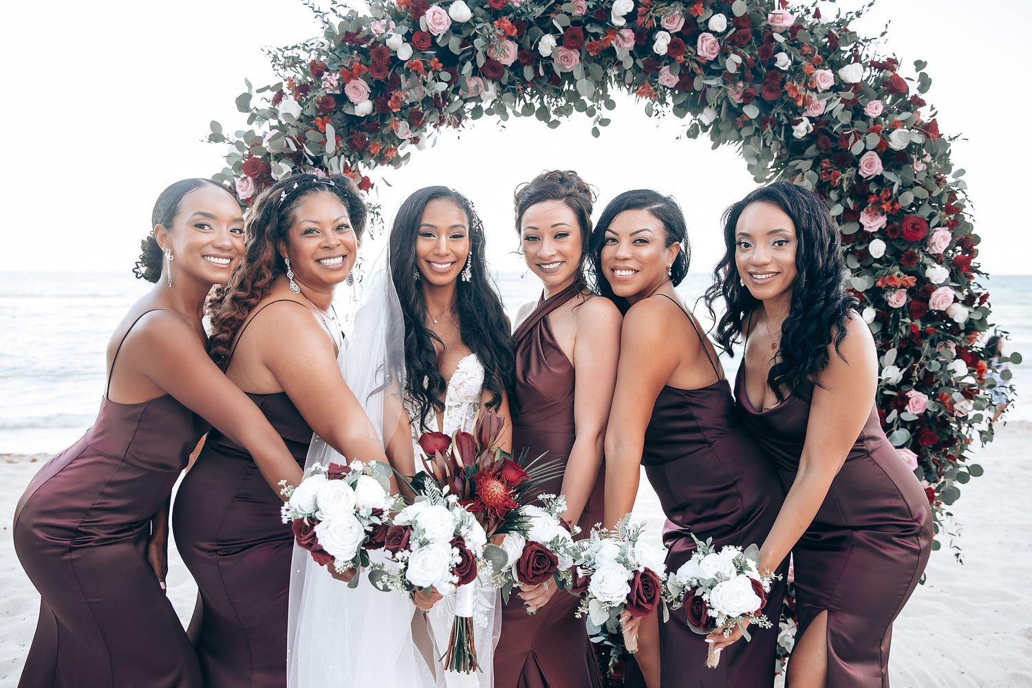 Bridesmaids in burgundy dresses pose by floral arch at beach wedding.