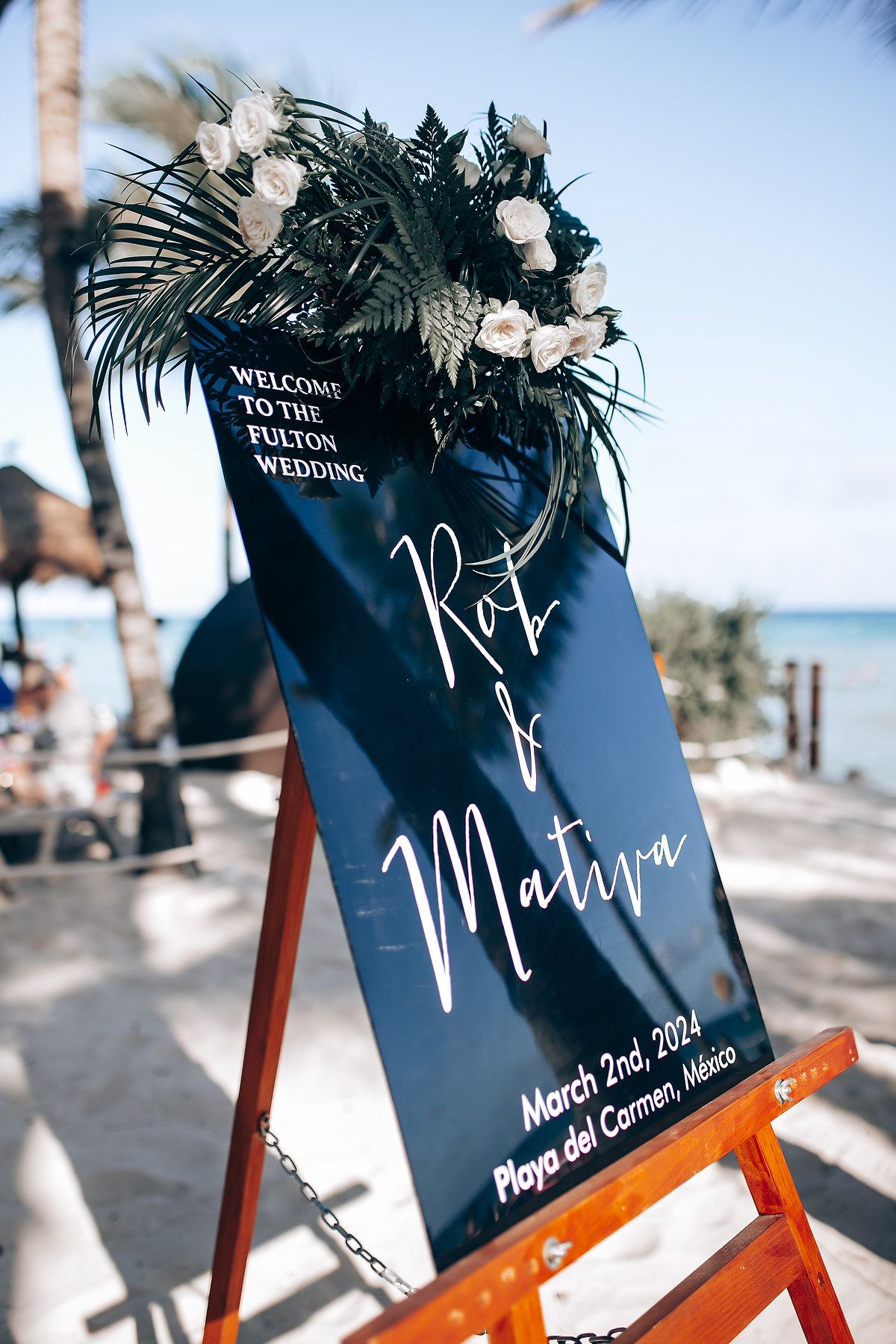 Navy blue wedding welcome sign with white florals displayed on wooden easel at beach venue.