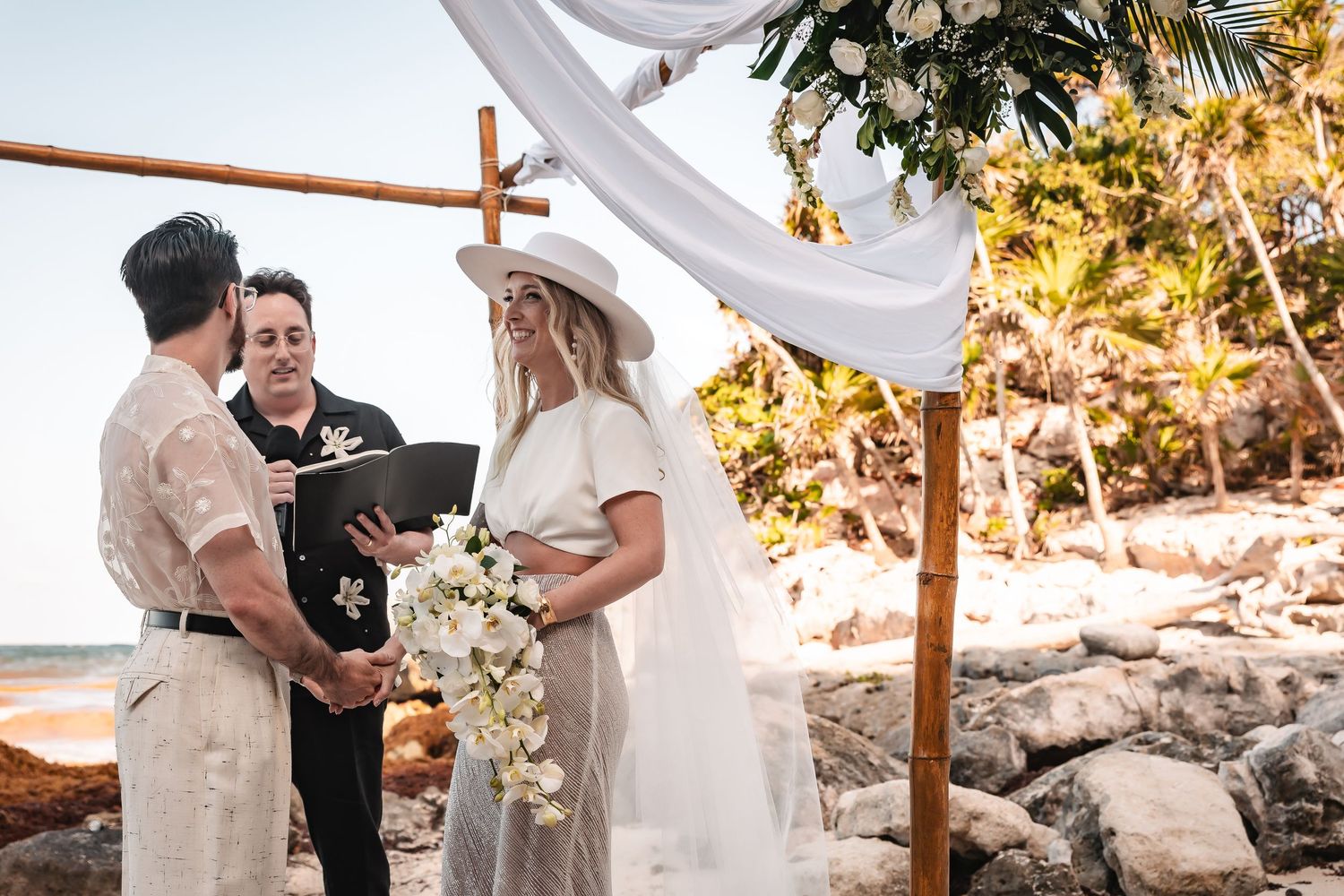 Intimate beach wedding ceremony with wooden cross altar and white fabric decor during golden hour.