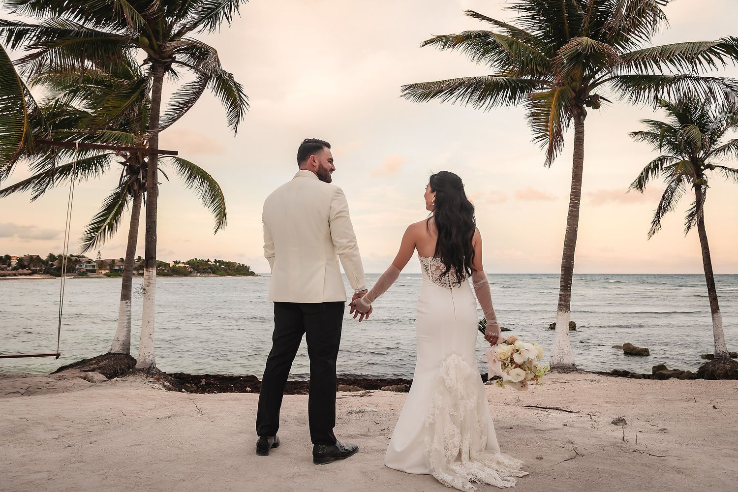 Couple holding hands on beach with palm trees and ocean view at sunset.