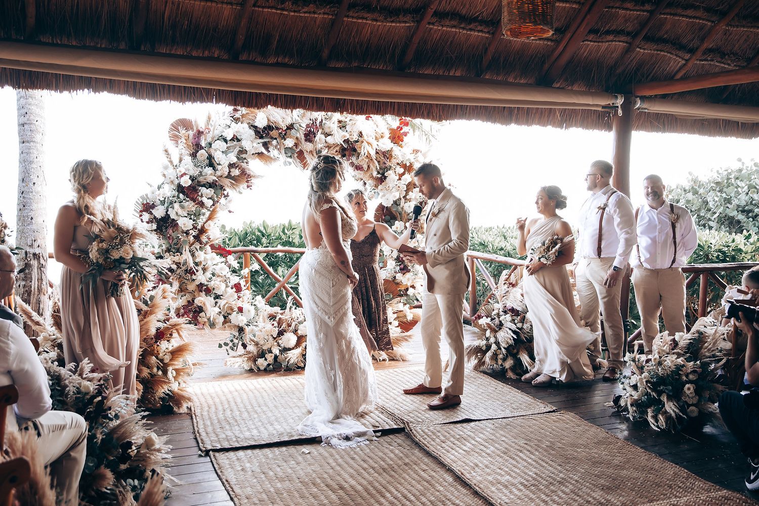 Panoramic view of a tropical wedding ceremony space with guests seated under a thatched roof pavilion.