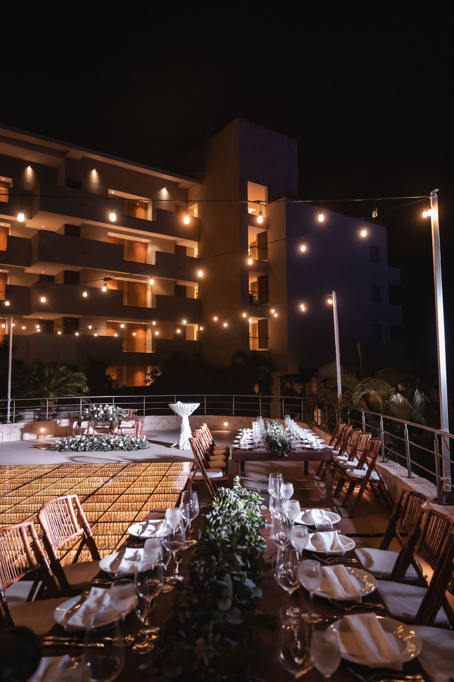 Elegant outdoor wedding reception setup with string lights and long tables on a terrace at night.